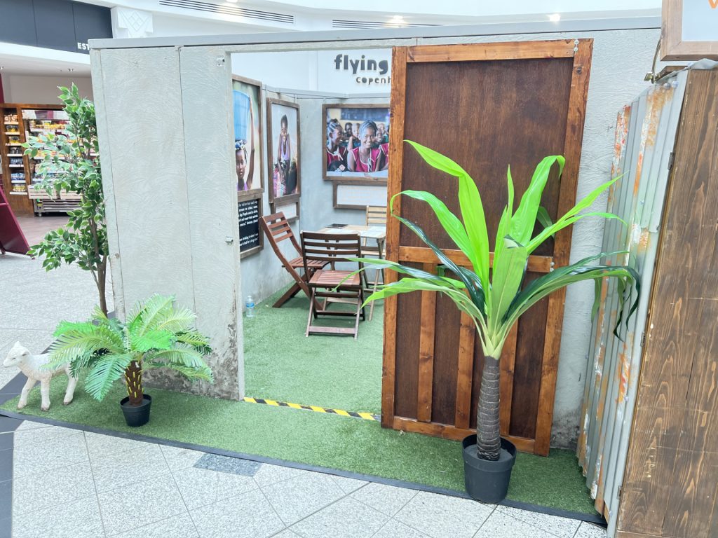 Indoor display booth in a mall with artificial grass floor, wooden panels, a small seating area, framed photos on the walls, and potted plants including a tall palm.