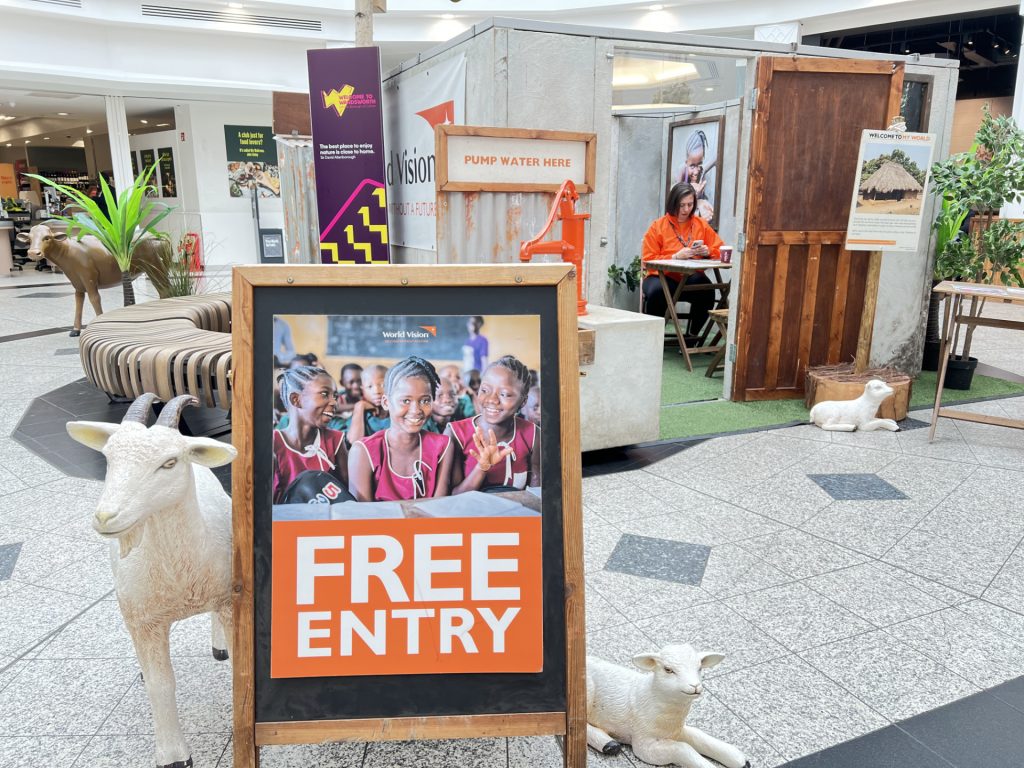 Promotional exhibit in a shopping mall with a “FREE ENTRY” sign, wooden frame, and life-size white sheep statues nearby.