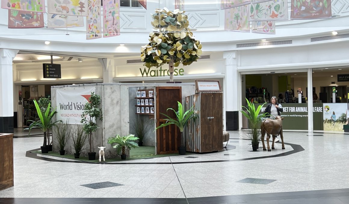 Indoor shopping-mall display: faux farm scene with potted plants, a small white goat statue, and a life-size brown horse statue near wooden panels and a World Vision banner.