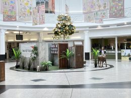 Indoor shopping-mall display: faux farm scene with potted plants, a small white goat statue, and a life-size brown horse statue near wooden panels and a World Vision banner.