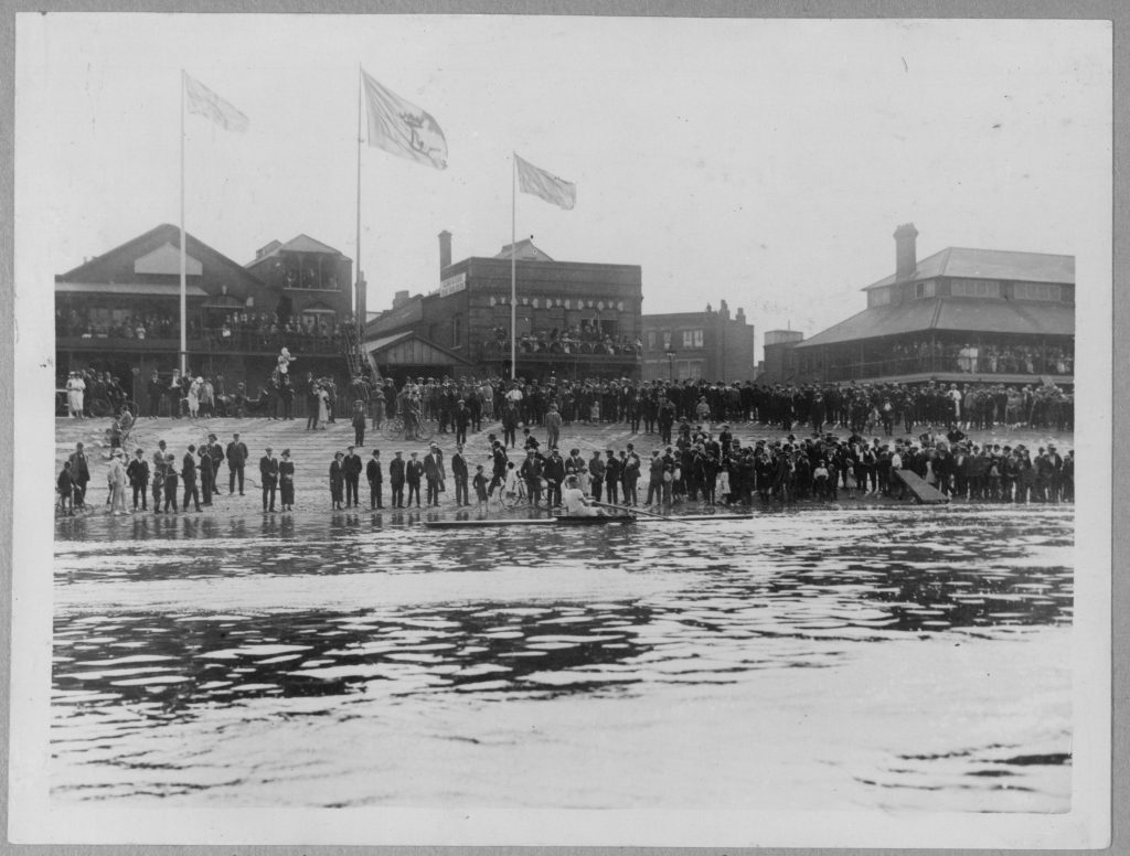 Black and white photograph of hundreds of spectators lining the Putney Embankment foreshore, with the industrial buildings of the north bank of the Thames visible across the river, 1925.