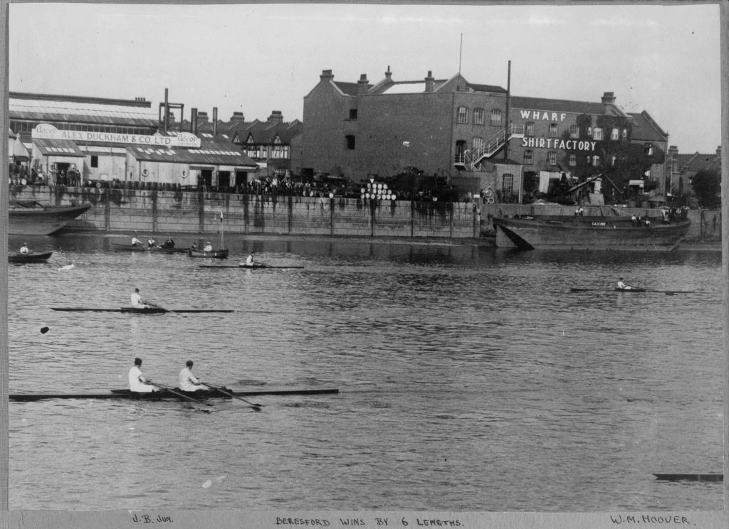Black and white photograph of two single scullers racing side by side on the Thames at Putney, with Alex Duckham & Co. and the Wharf Shirt Factory visible on the north bank. A handwritten caption, "Beresford wins by 6 lengths", appears at the bottom of the image.