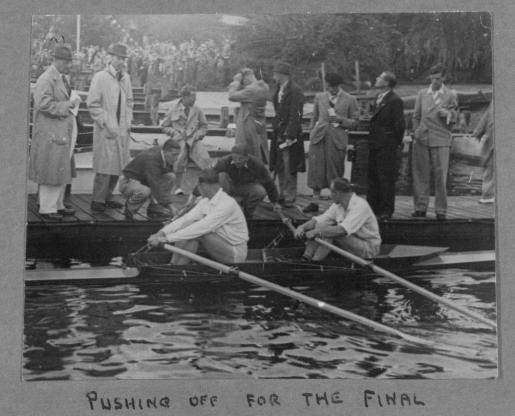 Black and white photograph of Jack Beresford and Dick Southwood seated low in their double scull at the dock, being pushed off by an attendant, with German officials in overcoats watching from the pontoon. Berlin, 1936.