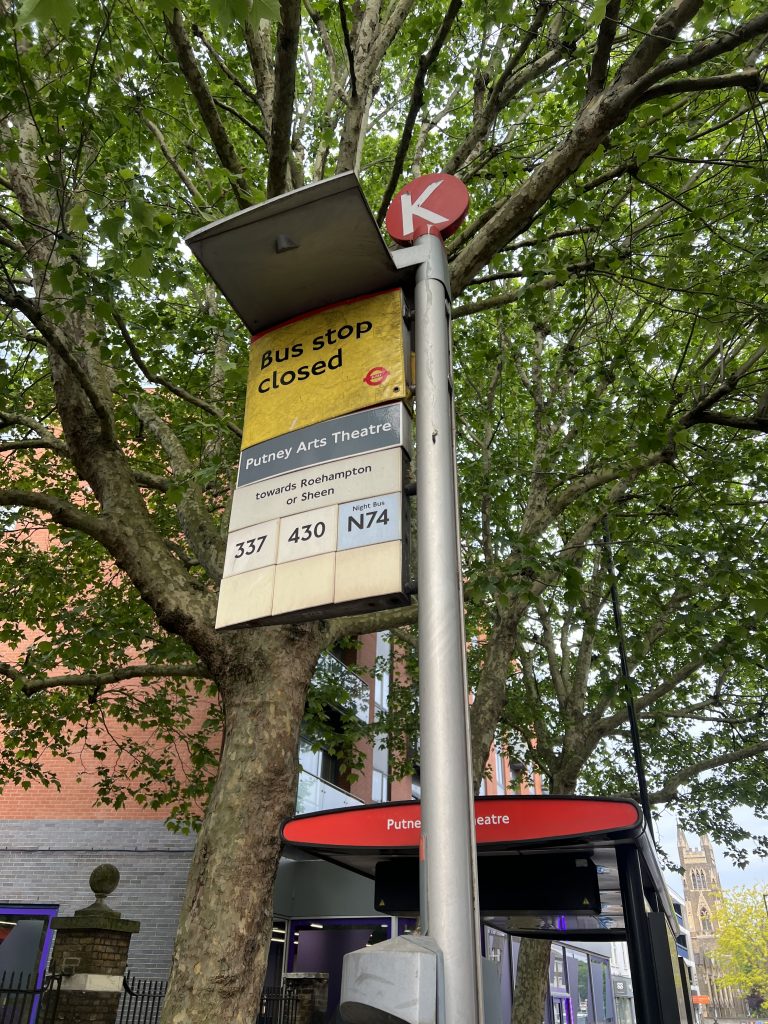 Bus stop sign mounted on a metal pole under green trees; yellow panel reads 'Bus stop closed' with 'Putney Arts Theatre' below and route numbers 337, 430, N74 visible.