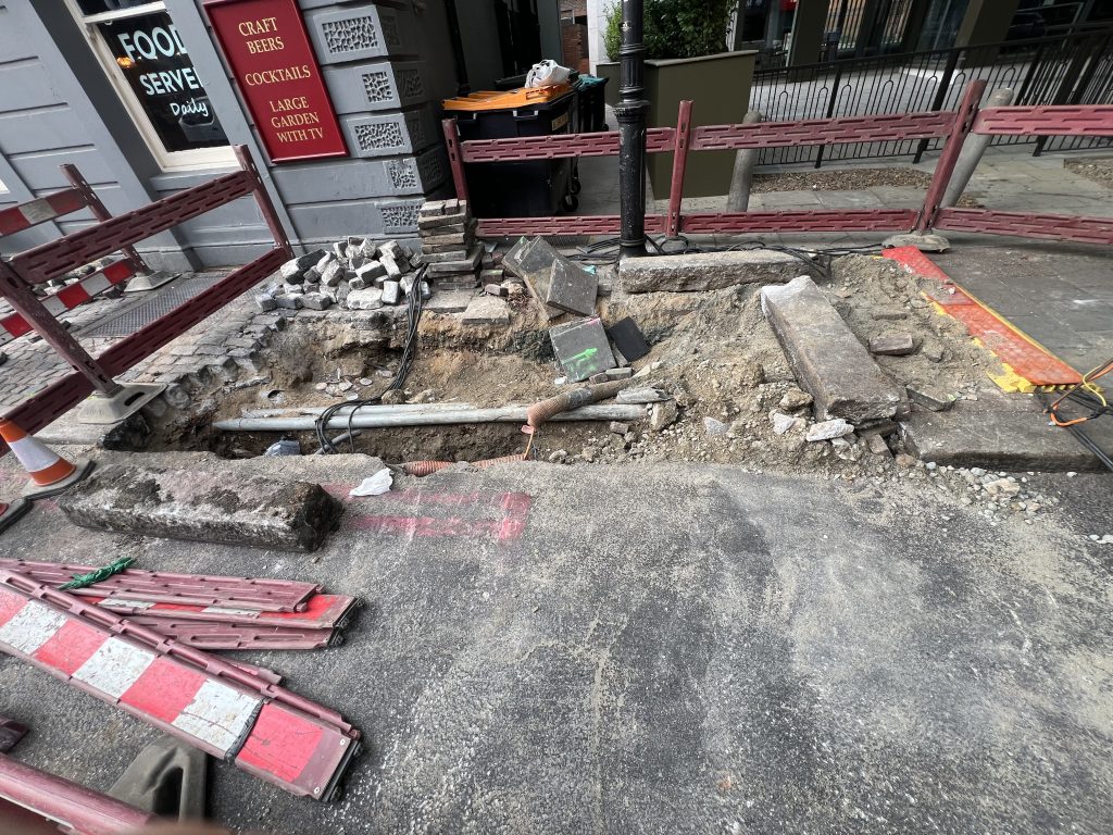 Construction work on a sidewalk with red plastic barricades, loose bricks, and exposed soil in front of a shop window.