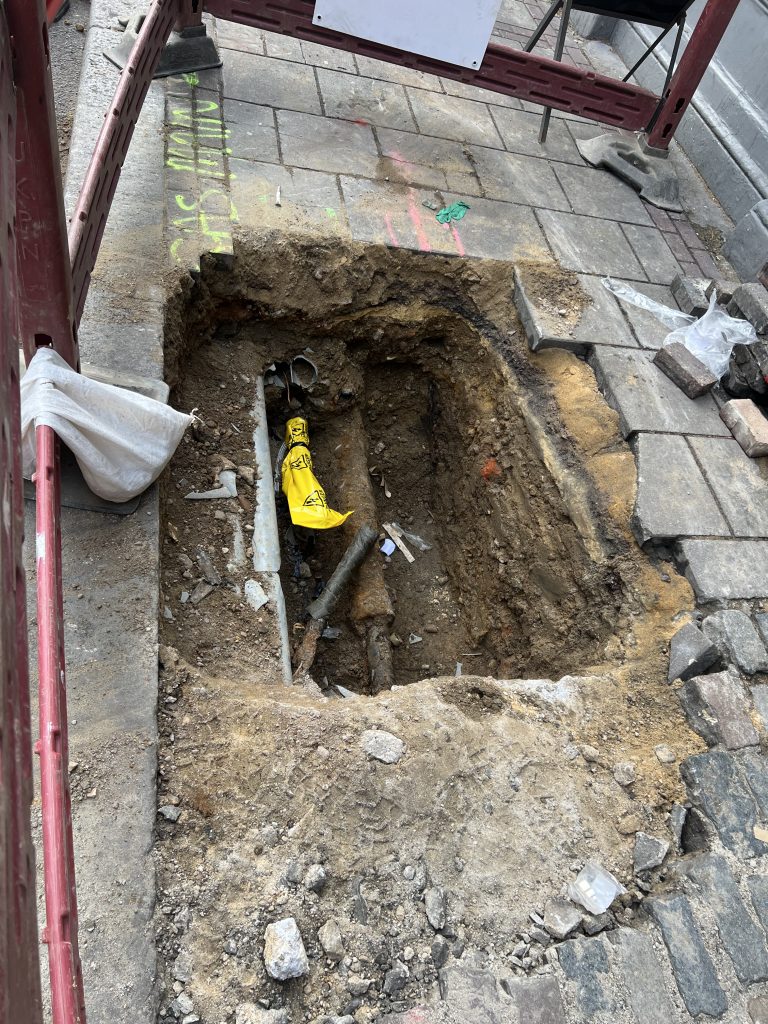 Open rectangular sidewalk trench with exposed soil and pipes, surrounded by loose bricks and construction barrier; a yellow warning bag lies inside the hole.