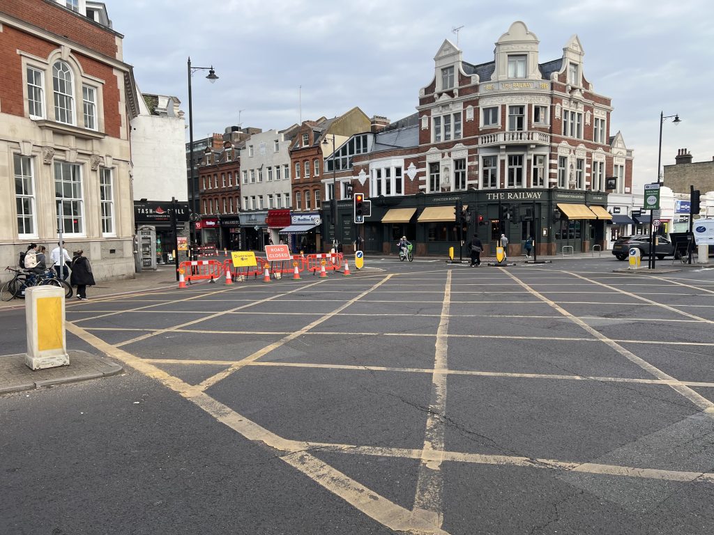 Urban street with historic red-brick building named The Railway on the corner and shopfronts along the road, pedestrians walking and cyclists nearby, and roadworks with orange barriers at center.