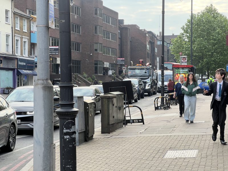 Pedestrians walking on a city sidewalk beside parked cars, brick office buildings, and a red bus on the street edge.