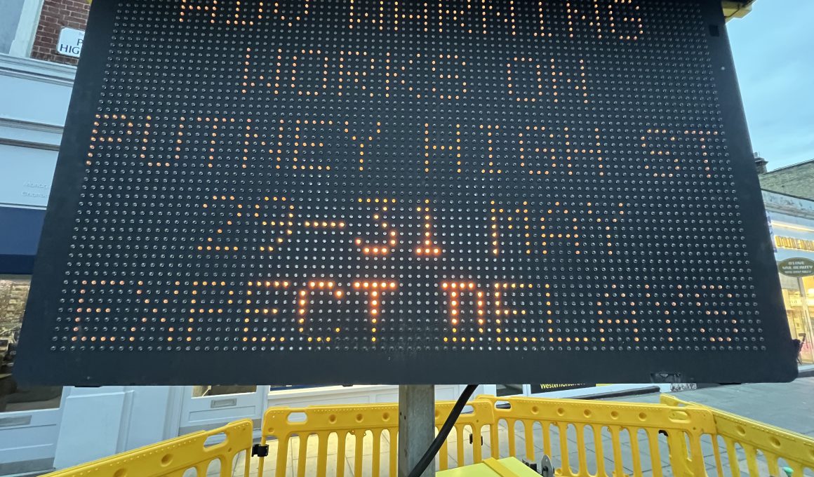 Tall black LED sign with orange dot-matrix text mounted on a pole at a construction site, surrounded by yellow safety barriers.