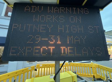 Tall black LED sign with orange dot-matrix text mounted on a pole at a construction site, surrounded by yellow safety barriers.