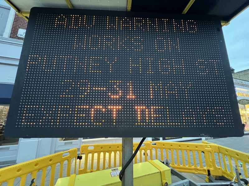 Tall black LED sign with orange dot-matrix text mounted on a pole at a construction site, surrounded by yellow safety barriers.