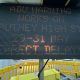 Tall black LED sign with orange dot-matrix text mounted on a pole at a construction site, surrounded by yellow safety barriers.