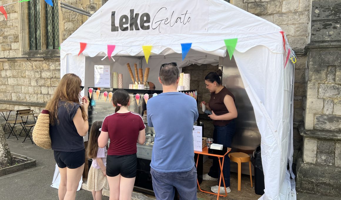 Customers line up at a white gelato booth under a stone building, with colorful pennant bunting and ice cream cones on display.