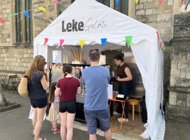 Customers line up at a white gelato booth under a stone building, with colorful pennant bunting and ice cream cones on display.