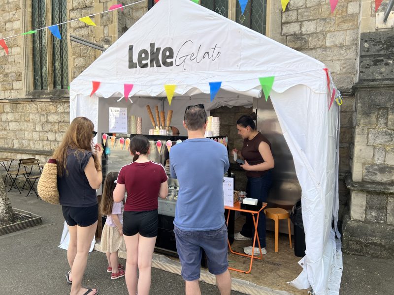 Customers line up at a white gelato booth under a stone building, with colorful pennant bunting and ice cream cones on display.