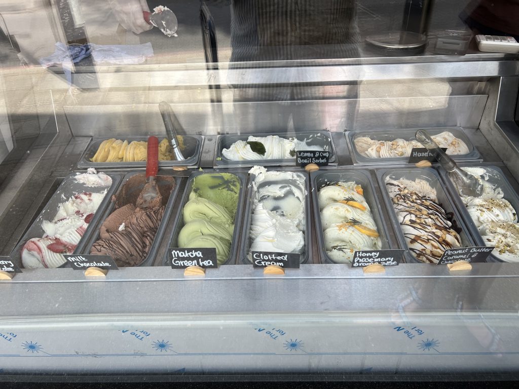 Assorted gelato flavors in a glass display case, labeled Milk Chocolate, Matcha Green Tea, Clotted Cream, Honeycomb, and Peanut Butter Caramel.