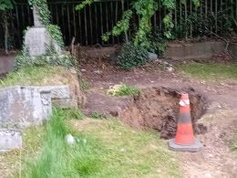 Blurry outdoor scene with ivy-covered stone structure on the left, a metal fence behind, dirt path, and an orange traffic cone in the foreground.