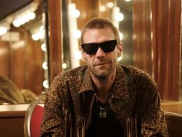 Man wearing leopard-print jacket and dark sunglasses sits in a dressing room with round mirror bulbs behind him, looking at the camera.