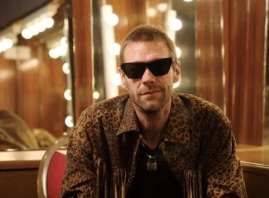 Man wearing leopard-print jacket and dark sunglasses sits in a dressing room with round mirror bulbs behind him, looking at the camera.