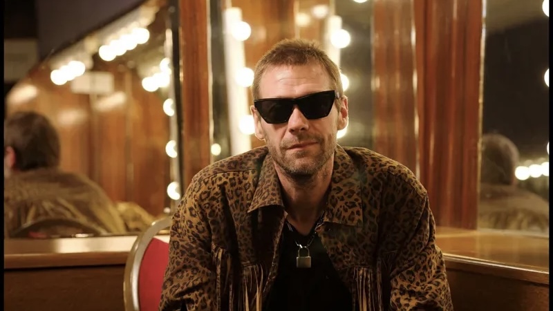 Man wearing leopard-print jacket and dark sunglasses sits in a dressing room with round mirror bulbs behind him, looking at the camera.