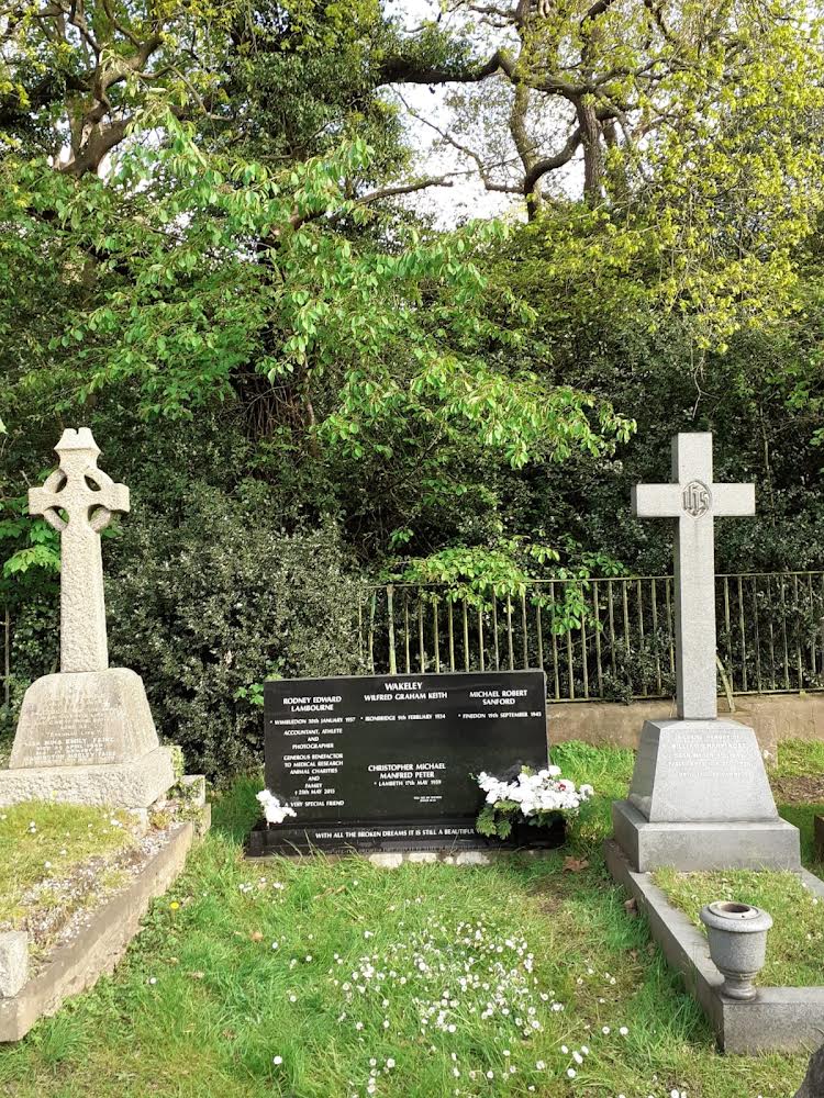 Cemetery scene with two stone crosses and a black memorial plaque surrounded by grass and trees in the background.