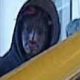 Person in a hooded jacket sitting by a window on a bus, holding a yellow handrail beside the passenger seat area.