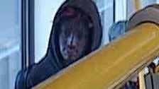 Person in a hooded jacket sitting by a window on a bus, holding a yellow handrail beside the passenger seat area.