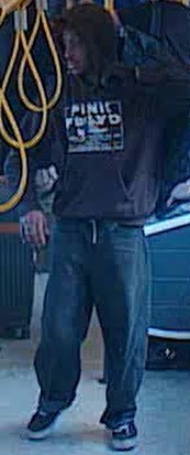 Man wearing a dark hoodie and jeans stands beside a blue car, with yellow hoses hanging overhead, suggesting a car wash setting.