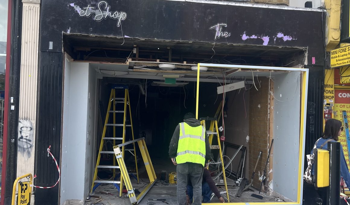 Front of a shop under renovation with workers in high‑visibility vests, ladders, and exposed interior wiring and debris outside a dark storefront.