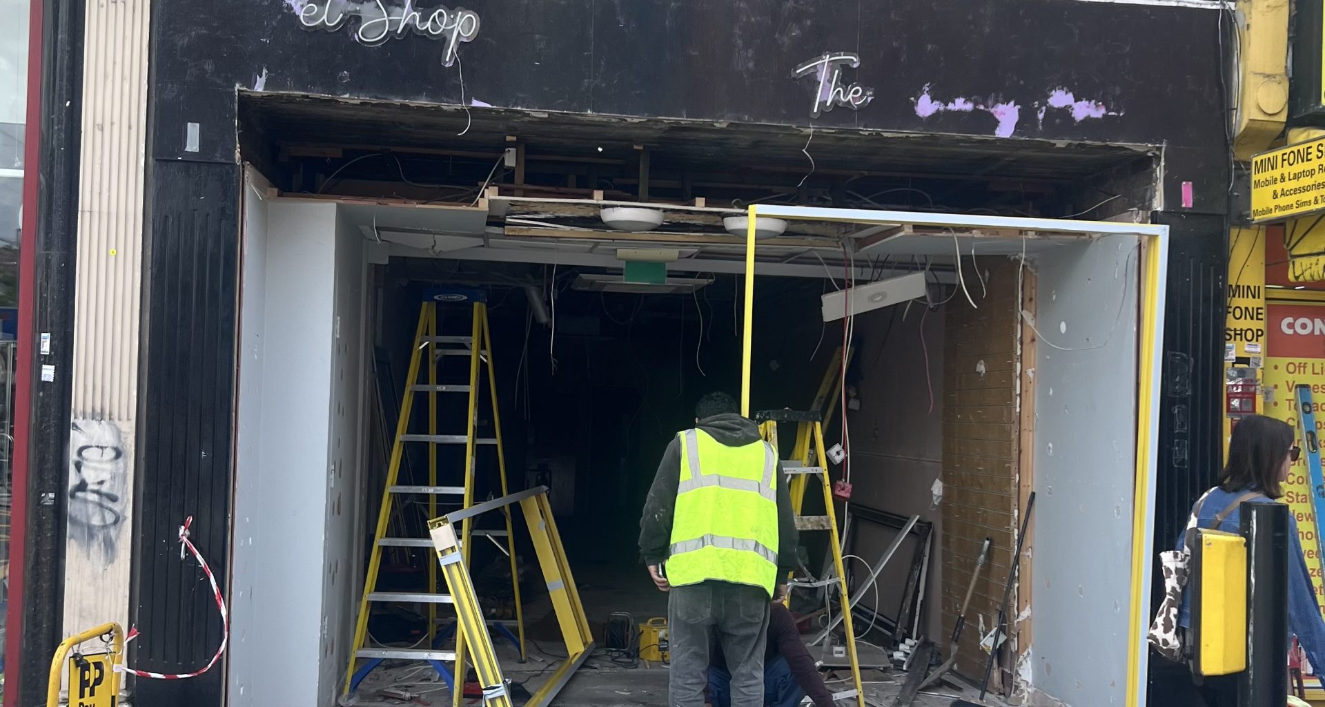 Front of a shop under renovation with workers in high‑visibility vests, ladders, and exposed interior wiring and debris outside a dark storefront.