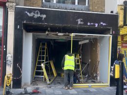 Front of a shop under renovation with workers in high‑visibility vests, ladders, and exposed interior wiring and debris outside a dark storefront.