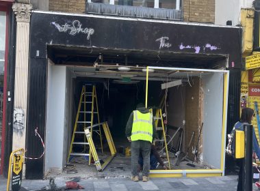 Front of a shop under renovation with workers in high‑visibility vests, ladders, and exposed interior wiring and debris outside a dark storefront.