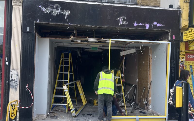 Front of a shop under renovation with workers in high‑visibility vests, ladders, and exposed interior wiring and debris outside a dark storefront.