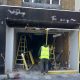 Front of a shop under renovation with workers in high‑visibility vests, ladders, and exposed interior wiring and debris outside a dark storefront.