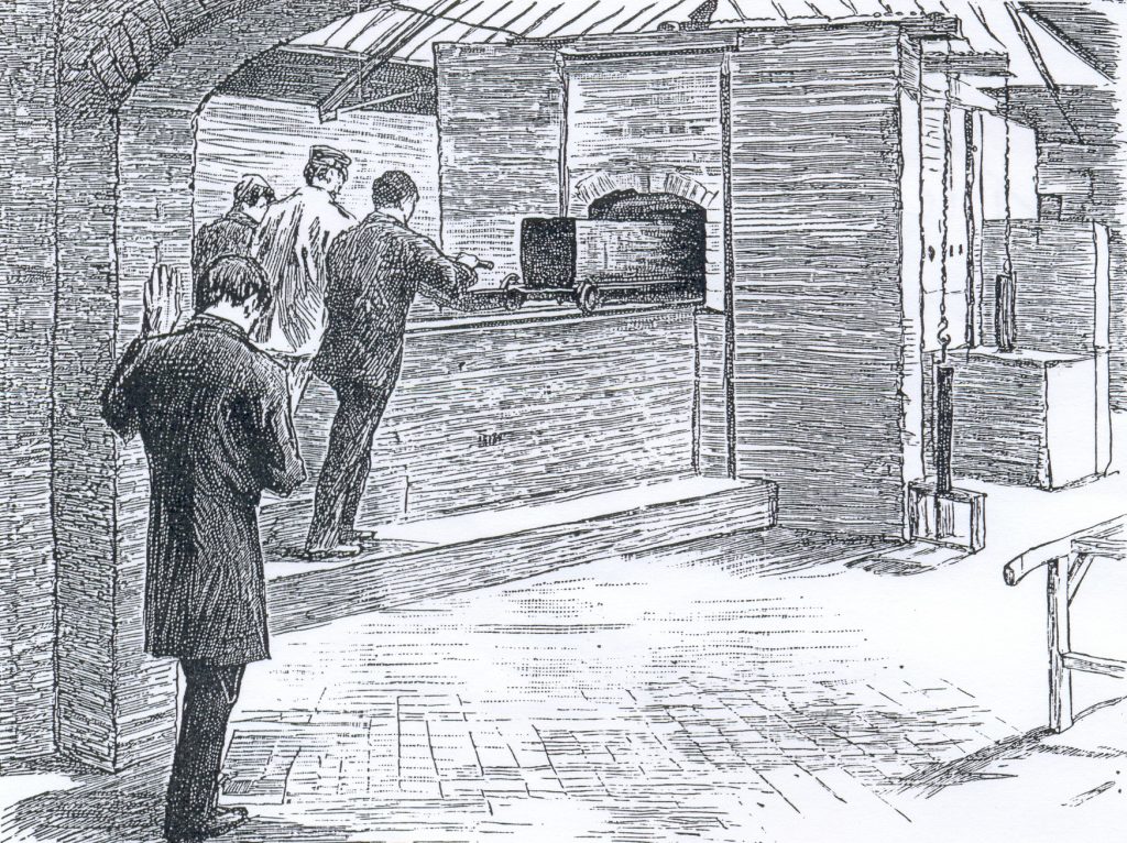 Three men stand at a long brick furnace in a workshop, one gesturing toward a cart with a large black oven on wheels behind a wooden barrier.