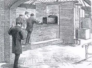 Three men stand at a long brick furnace in a workshop, one gesturing toward a cart with a large black oven on wheels behind a wooden barrier.