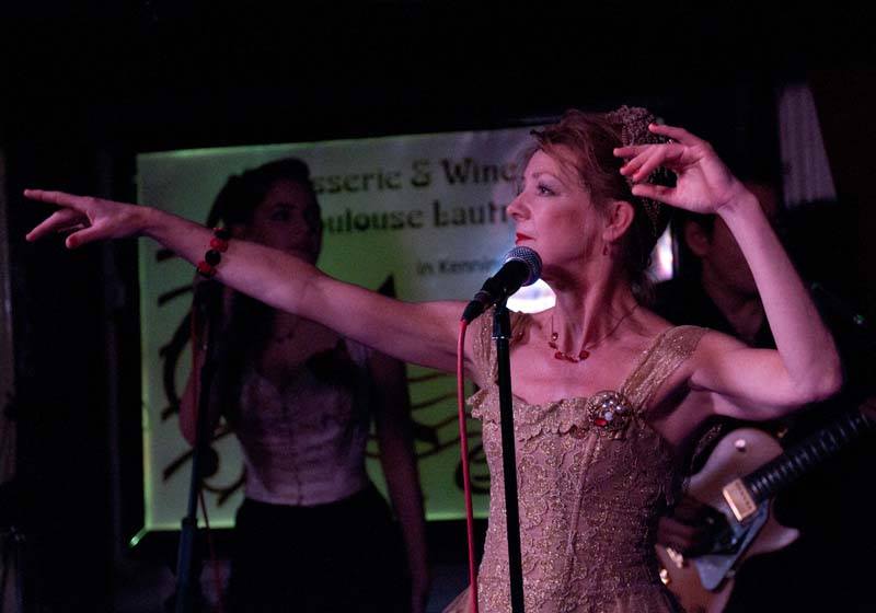 Female singer in a gold beaded dress performs on stage with arms outstretched and a microphone in front of her.
