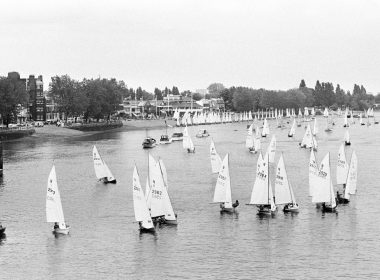 Many small sailboats with white sails on a wide river, with a tree-lined shore and buildings in the background.