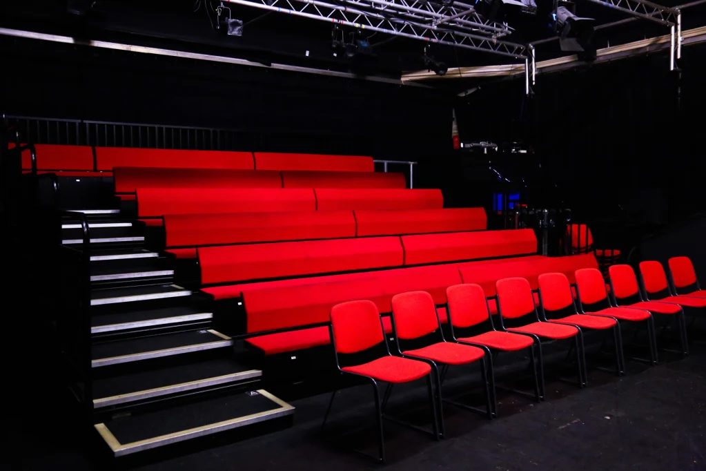 Red cushioned chairs arranged in tiered rows in a dark theater auditorium, empty and ready for an audience.