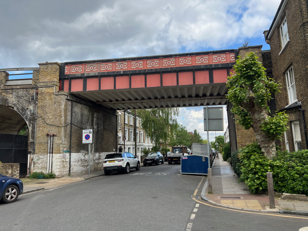 Urban street view beneath a decorative brick bridge with pink patterned panels, parked cars along the curb, and a tree on the right.