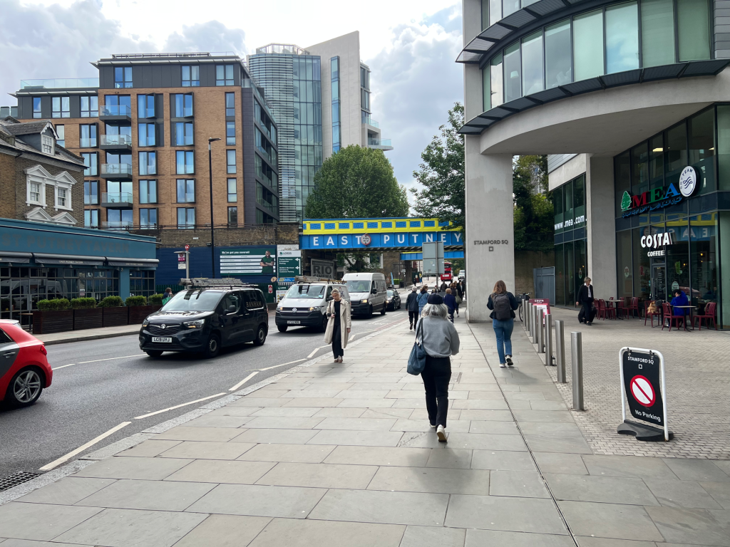 Pedestrians walk on a wide city sidewalk beside modern buildings and a blue East Putney bridge sign overhead.