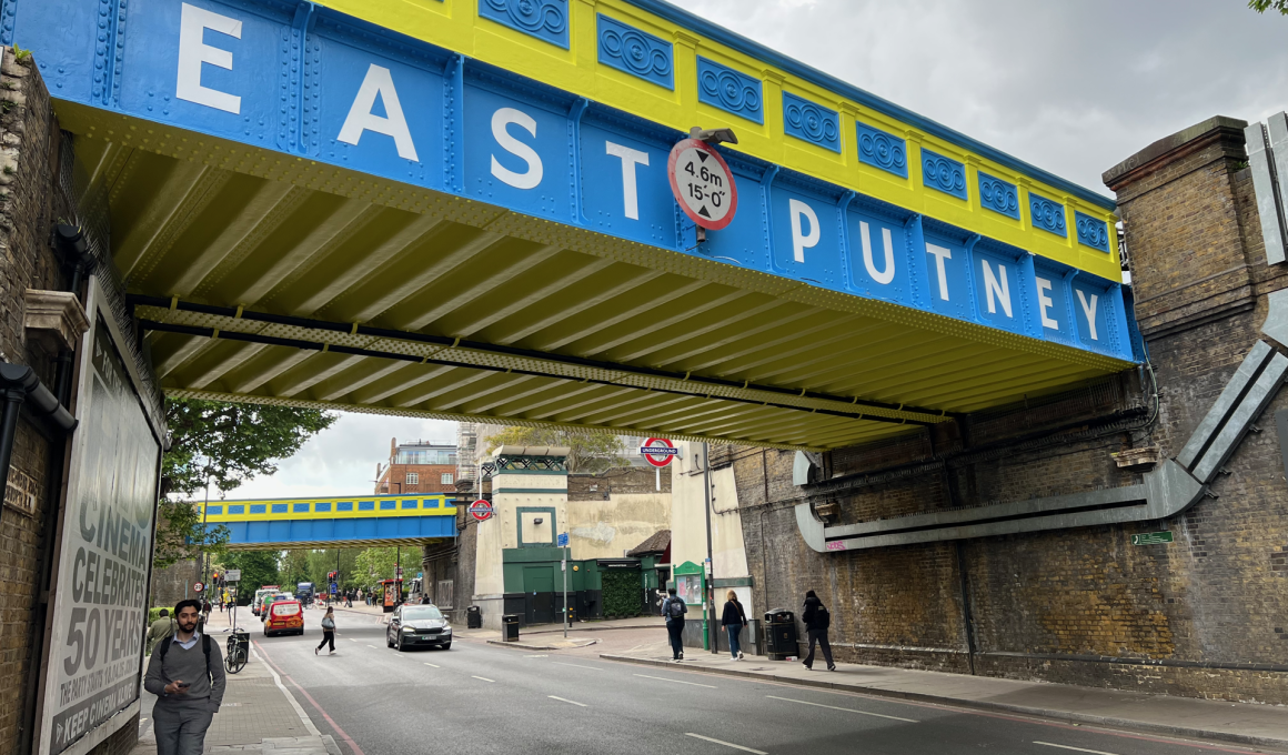 Underpass beneath a blue-and-yellow railway bridge with large white letters, pedestrians on the sidewalk.