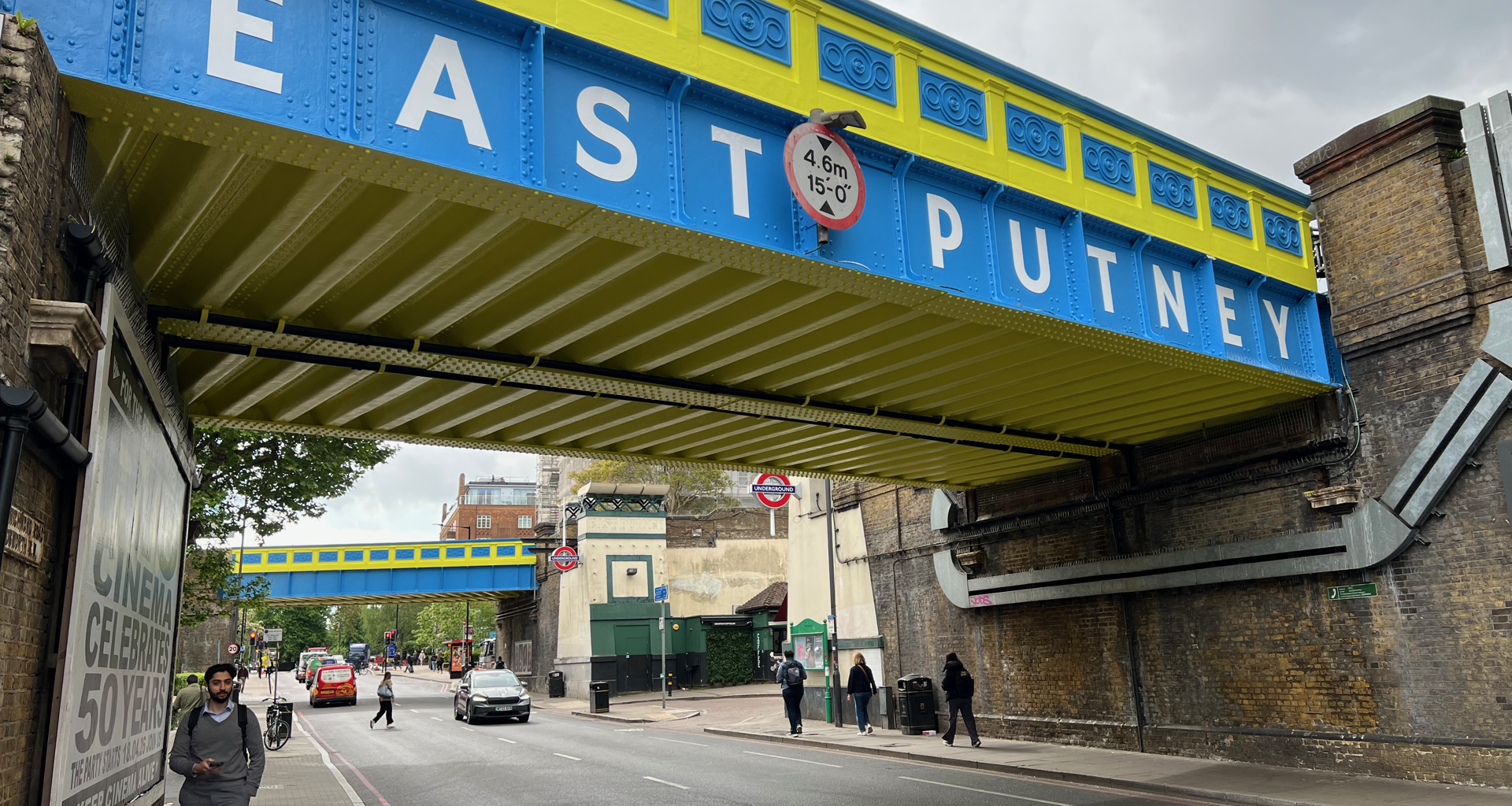 Underpass beneath a blue-and-yellow railway bridge with large white letters, pedestrians on the sidewalk.