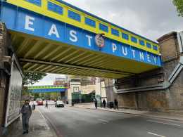 Underpass beneath a blue-and-yellow railway bridge with large white letters, pedestrians on the sidewalk.