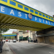 Underpass beneath a blue-and-yellow railway bridge with large white letters, pedestrians on the sidewalk.