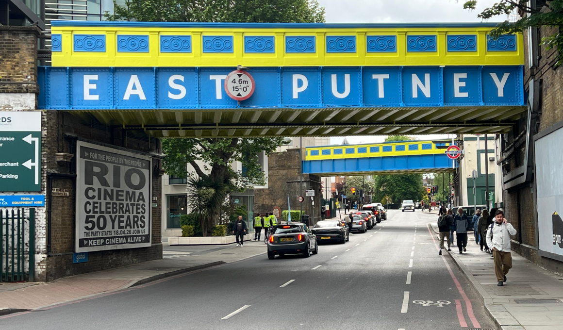 Colorful blue and yellow overhead bridge reading 'EAST PUTNEY' over a busy street with pedestrians and cars underneath.