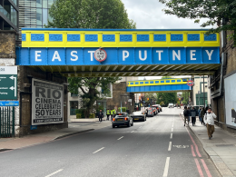 Colorful blue and yellow overhead bridge reading 'EAST PUTNEY' over a busy street with pedestrians and cars underneath.
