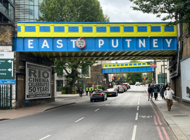 Colorful blue and yellow overhead bridge reading 'EAST PUTNEY' over a busy street with pedestrians and cars underneath.