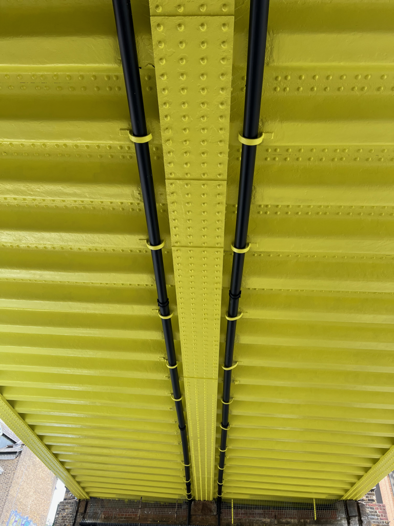 Bright yellow underside of a steel bridge with riveted beams and parallel black cables strapped along the center, visible brick buildings at the bottom edge.