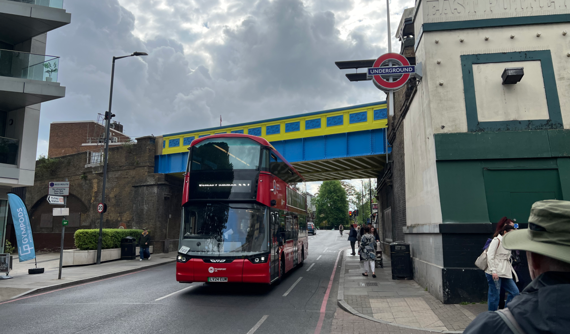 Red double-decker bus on a city street under a blue-and-yellow bridge, with a London Underground roundel sign nearby and pedestrians on the sidewalk.